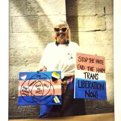A person with heart sunglasses holds signs advocating for trans liberation against a brick wall backdrop.