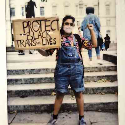 A person wearing a mask holds a sign saying "Protect Black Trans Lives," standing on steps.