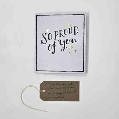 A white card, bordered in black with the text "SO PROUD of you" surrounded with gold stars. With a brown, handwritten luggage tag.