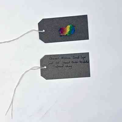 A piece of rainbow colour duct tape stuck to a brown luggage tag. With a second brown, handwritten luggage tag.