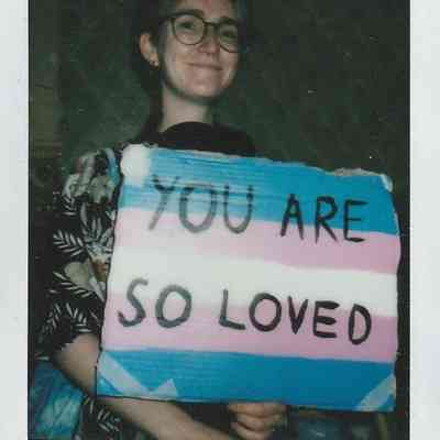 A person smiles while holding a colorful sign that reads, "You Are So Loved" in black letters.