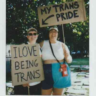 Two people are holding signs that express their pride and love for being transgender at an outdoor event.