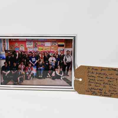 The image shows a group photo in a silver frame, with colourful protest signs hung on a pink wall at an exhibition. Next to the framed photograph is a brown tag with handwritten text on it.