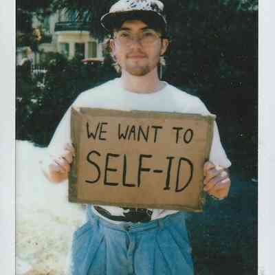 A person in glasses holds a cardboard sign saying “We want to self-ID” at an event.