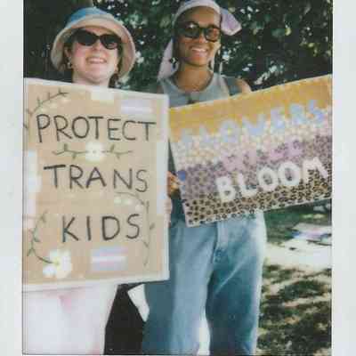 Two individuals are smiling and holding signs with supportive messages about trans rights, standing outdoors.