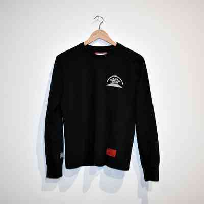 A black long-sleeve shirt is hanging on a wooden hanger against a plain wall. On the shirt, it says 'We Must Work To Protect Each Other' and 'Black Trans Lives Matter'.