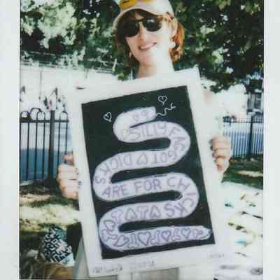 A person wearing sunglasses and a cap holds a colorful sign with playful text at an outdoor event.