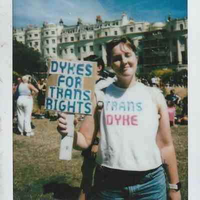 A person holds a sign reading "Dykes for Trans Rights," wearing a shirt with "Trans Dyke" printed on it.