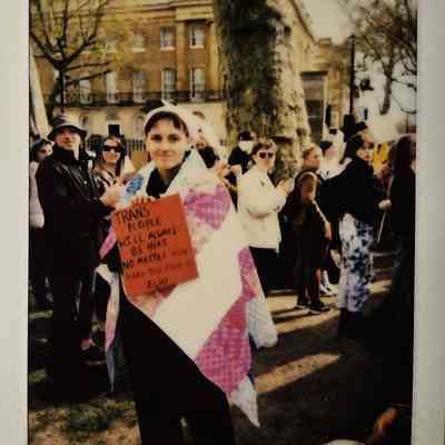 A person wrapped in a flag holds a sign at a gathering, surrounded by people and trees.