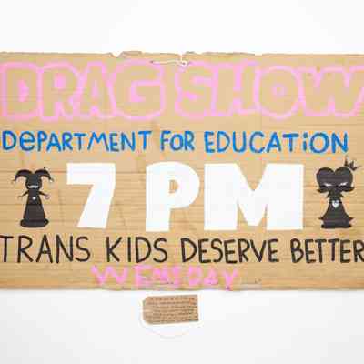 A hand drawn cardboard placard advertising a Drag Show a the Department for Education organised by Trans Kids Deserve Better. With a brown, handwritten luggage tag.