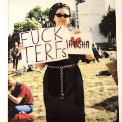 A person in sunglasses and a hat holds a sign which says 'FUCK TERFS' in a sunny outdoor setting.