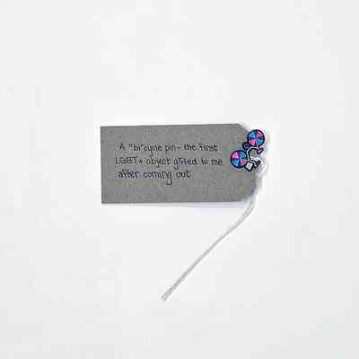 A pin of a bike which is in bisexual flag colours attached to a brown tag with writing on it.