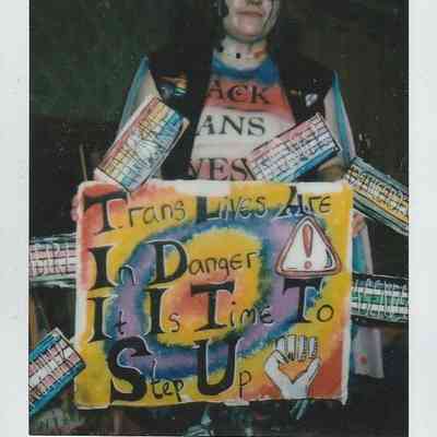 A person holds a colorful protest sign advocating for trans lives and action, with various badges displayed.