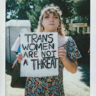 A person holds a sign reading "Trans women are not a threat" at an outdoor event.