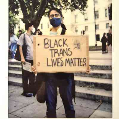 A masked individual holds a cardboard sign that reads "Black Trans Lives Matter" during a protest.