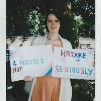 A person stands outdoors holding two signs, advocating for serious attention and housing, not just promises.