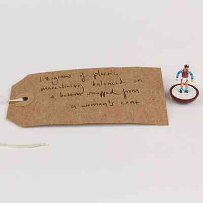A small figure stands on a button beside a handwritten tag describing plastic masculinity weight.