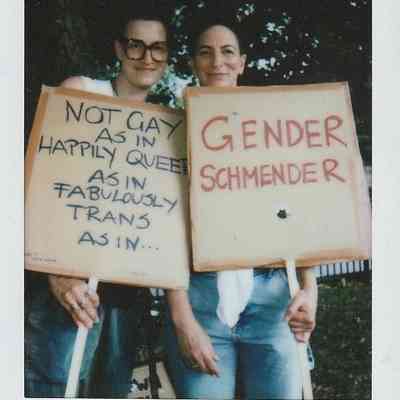 Two people hold signs with messages celebrating queer and transgender identities, standing confidently together outdoors.