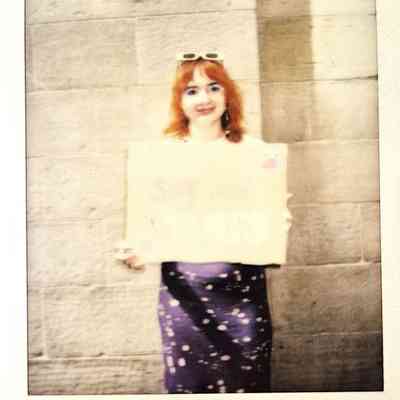 A polaroid of a person with red hair holds a partially visible sign while standing against a beige wall.
