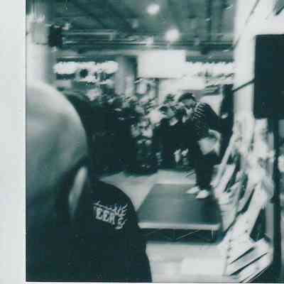 The image depicts a crowded indoor event with people gathered around a small stage in a dimly lit setting.