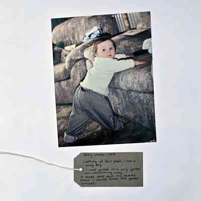 A photo of a young child with light brown hair wearing a white top and pin stripe trousers about to climb onto a sofa. With a brown handwritten luggage tag.
