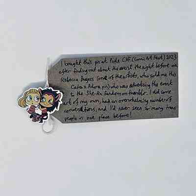 An enamel pin showing two people embracing and holding hands. One is blonde and wearing a red outfit and the other has brown hair and animal ears. They are characters from the cartoon She-Ra. The pin is attached to a brown tag with a handwritten message on it.