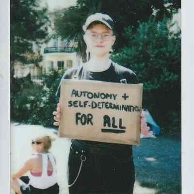 A person holds a cardboard sign stating "Autonomy + Self-Determination For All".