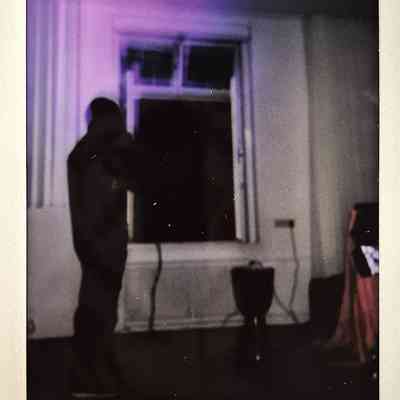 A silhouette stands by a window in a dimly lit room with purple light casting a subtle glow.