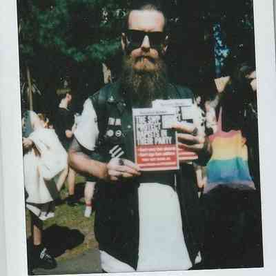 A bearded person in sunglasses holds a publication at an outdoor event, surrounded by people and trees.