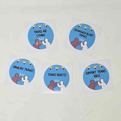 Five stickers on a white surface promote trans rights and support.