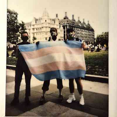 Three people hold a transgender pride flag outside near a large historic building.