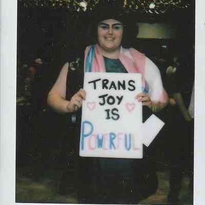 A person holds a sign saying "Trans Joy is Powerful," smiling under string lights.