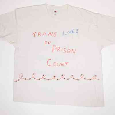 White T-shirt with handwritten text "TRANS LIVES IN PRISON COUNT" in red and blue and hand drawn red curly barbed wire with black 'x's.