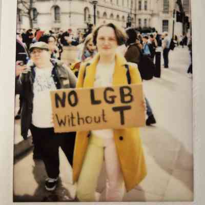 A person in a yellow coat holds a sign reading "No LGB without T" during a protest.