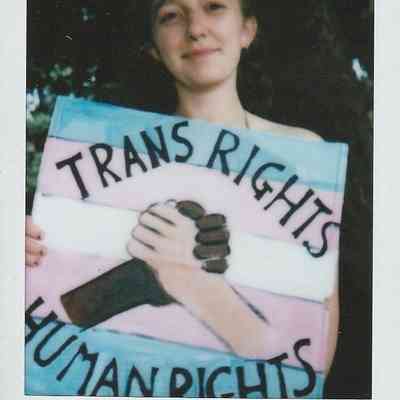 A person smiles, holding a sign that reads "Trans Rights Human Rights" with clasped hands.