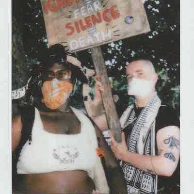 Two people, wearing masks, hold a sign that reads, "Ignorance + Fear + Silence = Death".