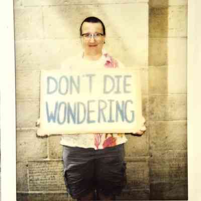 A polaroid of a person holds a sign reading "DON'T DIE WONDERING" against a stone wall backdrop.