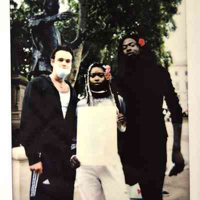 Three people stand together outdoors, two with flowers in their hair, in front of a statue.