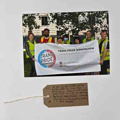 A group poses with a "Trans Pride Birmingham" banner, smiling and wearing yellow vests.