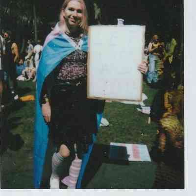 A person draped in a colorful flag holds a sign stating "Let Us Compete".