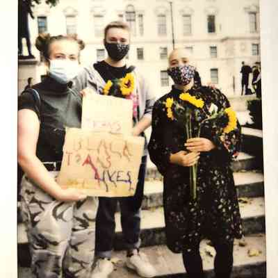 Three people wearing masks stand together holding sunflowers and a sign advocating for the protection of trans Black lives.
