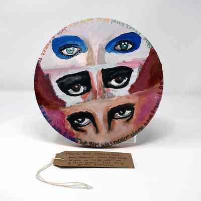 The circular artwork features three painted eye pairs with handwritten text surrounding them. With a brown handwritten luggage tag.
