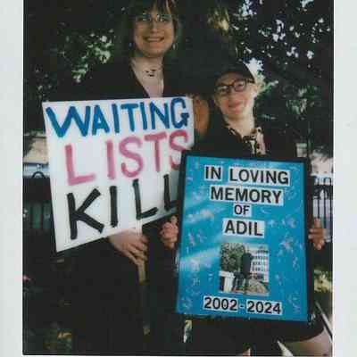Two individuals hold signs advocating for healthcare reform, one reads "Waiting Lists Kill," urging change.