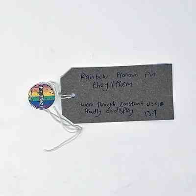 The image shows a rainbow pronoun pin labeled "they/them" with a tag describing its usage