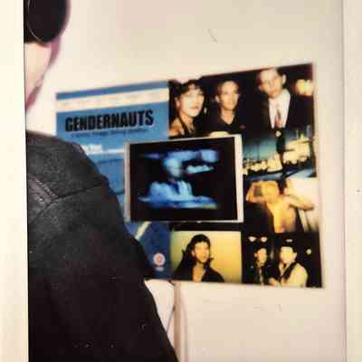 A person stands in front of a wall displaying posters and photographs, with a focus on "Gendernauts".