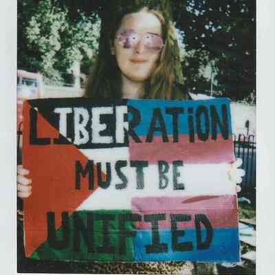 A person holds a colorful sign that reads "Liberation must be unified" beneath a tree.