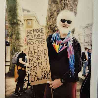 A person stands holding a protest sign which says "NO MORE TORTURE NO ONE LEFT BEHIND BORIS DON'T BETRAY US YOU COWARD".