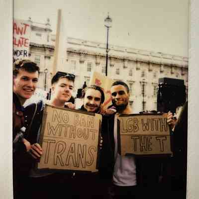 Four people are standing together, holding signs which says "NO BAN WITHOUT TRANS" and "LGB WITH THE T".