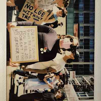 People are participating in an outdoor protest and one person is holding a sign which says "DEAR TORIES, JUST BECAUSE YOU ARE 2D DOESN'T MEAN MY GENDER IS".