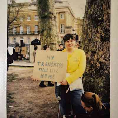 A person in a yellow shirt holds a sign reading "My transness feels like home".